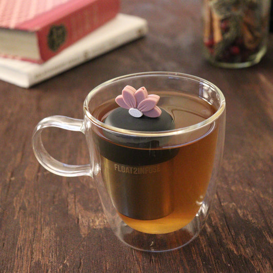 pink lotus flower tea infuser for loose leaf tea with a fine mesh stainless steel tea infuser that reads FLOAT2INFUSE on a black base floating in a glass tea mug with books and loose leaf tea in the background