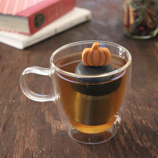 orange pumpkin tea infuser for loose leaf tea with a fine mesh stainless steel tea infuser that reads FLOAT2INFUSE on a black base floating in a glass tea mug with books and loose leaf tea in the background