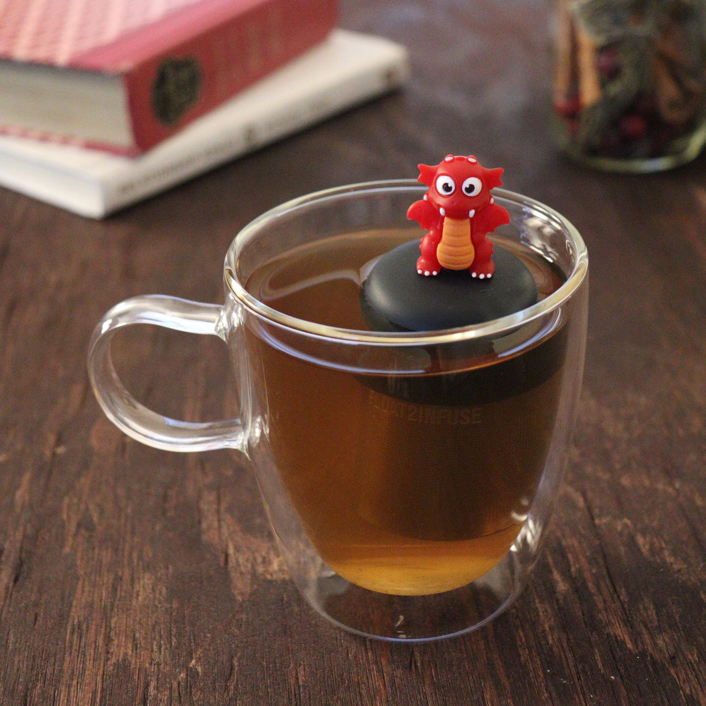 red dragon animal tea infuser for loose leaf tea with a fine mesh stainless steel tea infuser that reads FLOAT2INFUSE on a black base floating in a glass tea mug with books and loose leaf tea in the background