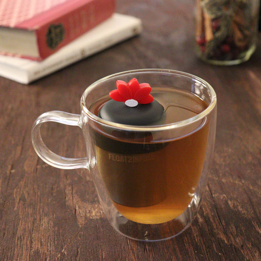 red lotus flower tea infuser for loose leaf tea with a fine mesh stainless steel tea infuser that reads FLOAT2INFUSE on a black base floating in a glass tea mug with books and loose leaf tea in the background