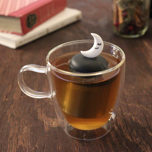 smiling crescent moon tea infuser for loose leaf tea with a fine mesh stainless steel tea infuser that reads FLOAT2INFUSE on a black base floating in a glass tea mug with books and loose leaf tea in the background