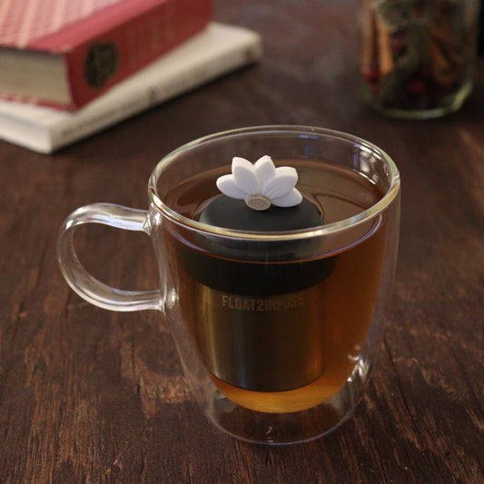 white lotus flower tea infuser for loose leaf tea with a fine mesh stainless steel tea infuser that reads FLOAT2INFUSE on a black base floating in a glass tea mug with books and loose leaf tea in the background