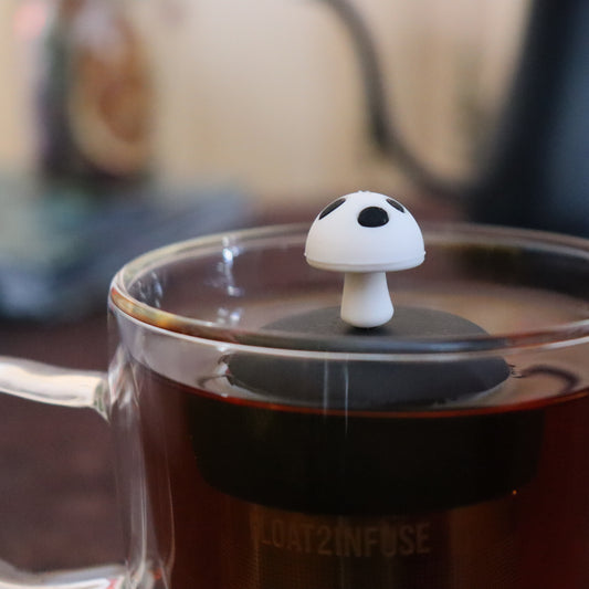 white and black mushroom tea infuser for loose leaf tea with a fine mesh stainless steel strainer that reads FLOAT2INFUSE on a black base floating in a glass tea mug with tea kettle in the background