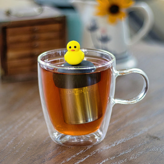 yellow rubber duck tea infuser for loose leaf tea with a fine mesh stainless steel tea strainer that reads FLOAT2INFUSE on a black base floating in a glass tea mug with a drink pitcher in the background