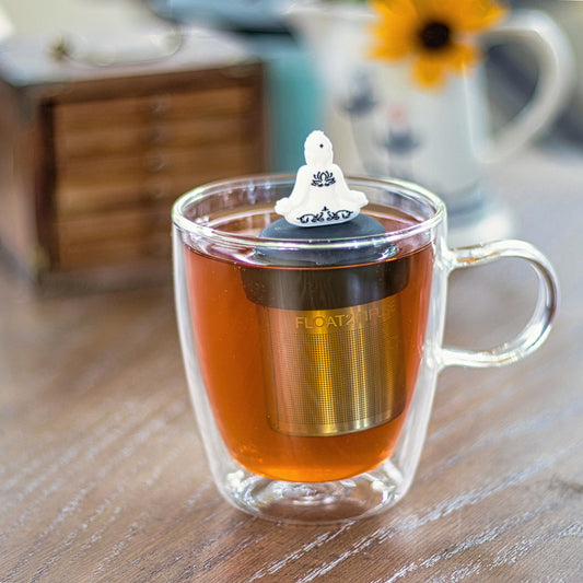 white yogi tea infuser for loose leaf tea with a fine mesh stainless steel strainer that reads FLOAT2INFUSE on a black basefloating in a glass tea mug with a water pitcher in the background