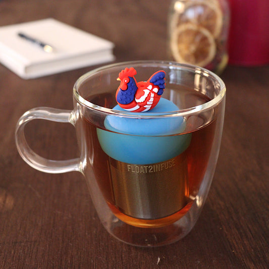 Red, white, and blue patterned chicken tea infuser for loose leaf tea with a fine mesh stainless steel tea strainer
that reads FLOAT2INFUSE on a blue base with a pen laying across a book and dried orange slices in a mason jar in the background