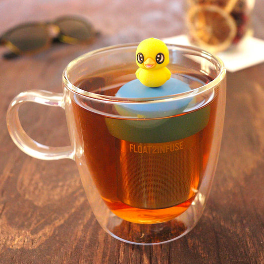 Baby duck animal tea infuser for loose leaf tea with a fine mesh stainless steel tea infuser that reads FLOAT2INFUSE on a blue base floating in a glass tea mug with loose leaf tea and sunglasses in the background