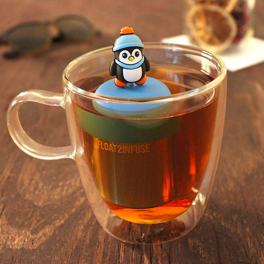 cute animal penguin wearing a blue hat and scarf tea infuser for loose leaf tea with a fine mesh stainless steel tea infuser that reads FLOAT2INFUSE on a blue base floating in a glass tea mug with loose leaf tea and sunglasses in the background
