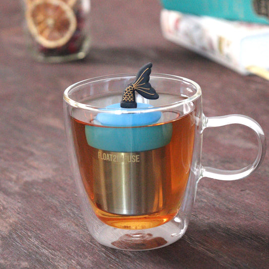 black and gold mermaid tale tea infuser for loose leaf tea on a blue base with a fine mesh stainless steel tea strainer that reads FLOAT2INFUSE floating in a glass tea mug with loose leaf tea and books in the background