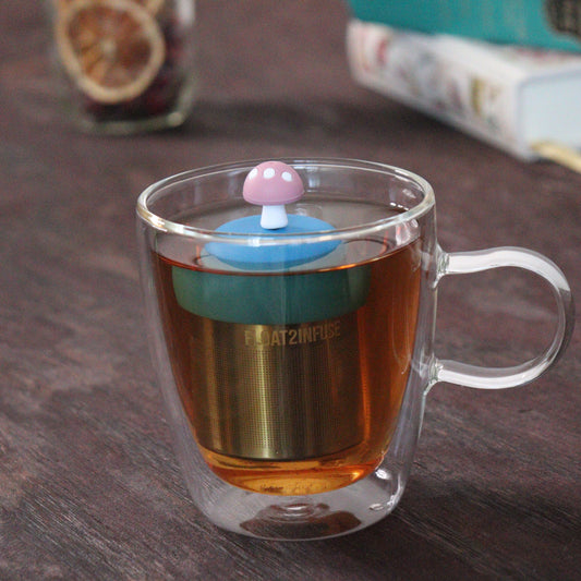 pink mushroom tea infuser for loose leaf tea with a fine mesh stainless steel tea strainer that reads FLOAT2INFUSE on a blue base floating in a glass tea mug with loose leaf tea and books in the background