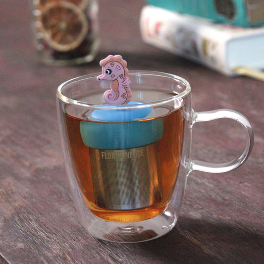 pink seahorse animal tea infuser for loose leaf tea with a fine mesh stainless steel tea strainer that reads FLOAT2INFUSE on a blue base floating in a glass tea mug with loose leaf tea and books in the background