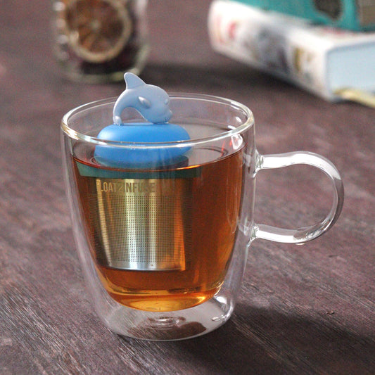 grey dolphin animal tea infuser for loose leaf tea on a blue base with a fine mesh stainless steel tea strainer that reads FLOAT2INFUSE floating in a glass tea mug with loose leaf tea and books in the background
