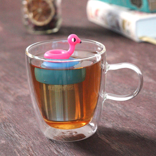 cute pink flamingo pool float tea infuser for loose leaf tea on a blue base with a fine mesh stainless steel tea strainer that reads FLOAT2INFUSE floating in a glass tea mug with loose leaf tea and books in the background