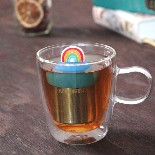 rainbow tea infuser for loose leaf tea with a fine mesh stainless steel tea strainer that reads FLOAT2INFUSE on a blue base floating in a glass tea mug with loose leaf tea and books in the background