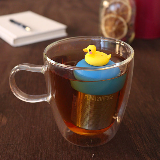 Yellow rubber duckie tea infuser for loose leaf tea with a fine mesh stainless steel tea strainer
that reads FLOAT2INFUSE on a blue base with a book with a pen and dried orange slices in a mason jar in the background
