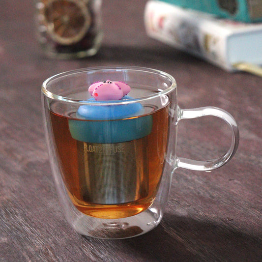 cute pink starfish animal float tea infuser for loose leaf tea on a blue base with a fine mesh stainless steel tea strainer that reads FLOAT2INFUSE floating in a glass tea mug with loose leaf tea and books in the background