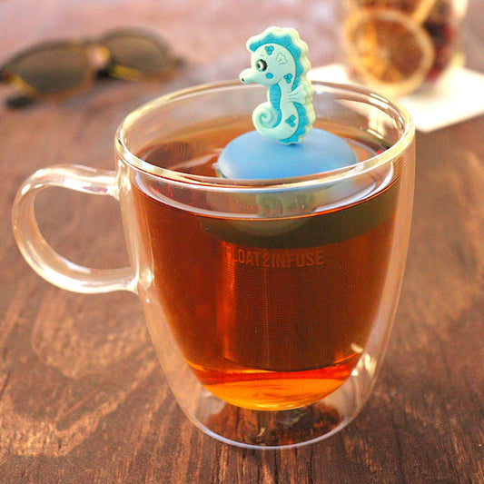 teal seahorse tea infuser for loose leaf tea with a fine mesh stainless steel tea infuser that reads FLOAT2INFUSE on a blue base floating in a glass tea mug with loose leaf tea and sunglasses in the background