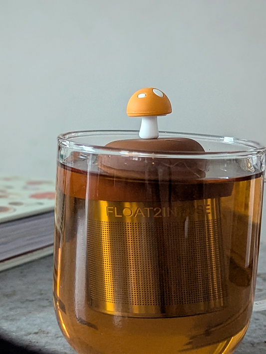 amber yellow mushroom with white dots tea infuser for loose leaf tea with a fine mesh stainless steel tea strainer that reads FLOAT2INFUSE on a brown base floating in a glass tea mug with a book in the background