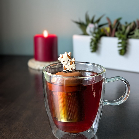 Brown and beige goat tea infuser for loose leaf tea with a fine mesh stainless steel tea strainer that reads FLOAT2INFUSE on a brown base with plants and a candle in the background