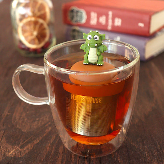 dark green dragon tea infuser for loose leaf tea with a fine mesh stainless steel tea strainer that reads FLOAT2INFUSE on a brown base floating in a glass tea mug with loose leaf tea and books in the background