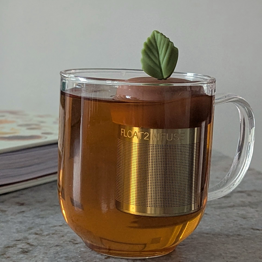 green herb leaf tea infuser for loose leaf tea with a fine mesh stainless steel tea steeper that reads FLOAT2INFUSE on a brown base floating in a glass tea mug with books in the background