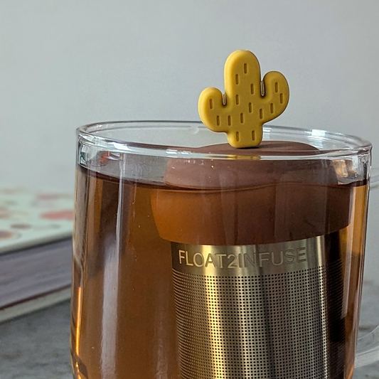 yellow cactus tea infuser for loose leaf tea with a fine mesh stainless steel tea steeper that reads FLOAT2INFUSE on a brown base floating in a glass tea mug with books in the background