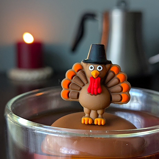 Thanksgiving turkey wearing a black pilgrim top hat tea infuser for loose leaf tea with a fine mesh stainless steel tea strainer that reads FLOAT2INFUSE on a brown base with a lit candle and a tea kettle in the background
