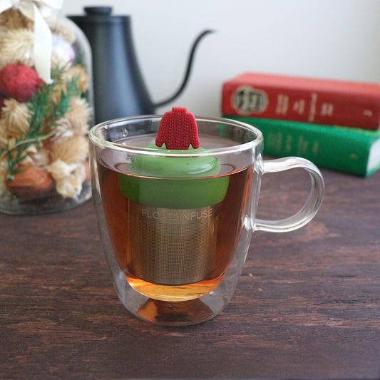 red knit sweater for loose leaf tea with fine mesh stainless steel tea strainer that reads FLOAT2INFUSE on a dark green base with books, a tea kettle, and christmas decorations in the background
