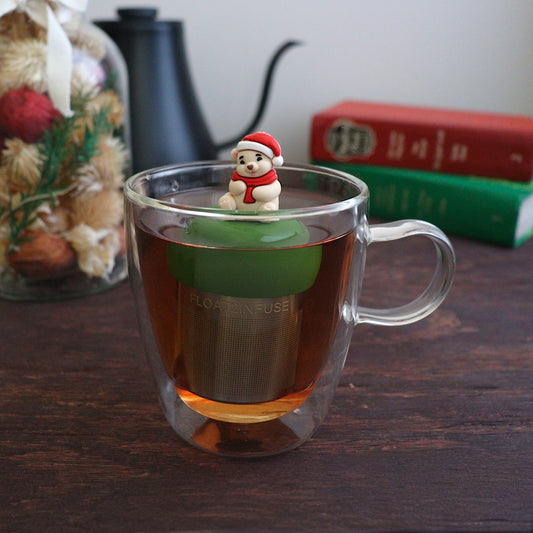 Cozy white polar bear wearing a red and white santa hat and a red scarf tea infuser for loose leaf tea with a fine mesh stainless steel tea strainer
that reads FLOAT2INFUSE on a dark green base with books, a tea kettle, and christmas decorations in the background