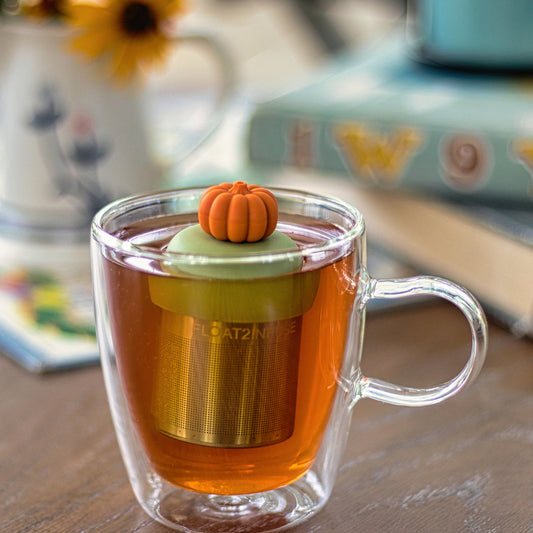 3D orange pumpkin tea infuser for loose leaf tea with a fine mesh stainless steel tea strainer that reads FLOAT2INFUSE on a green base floating in a glass tea mug
with books in the background