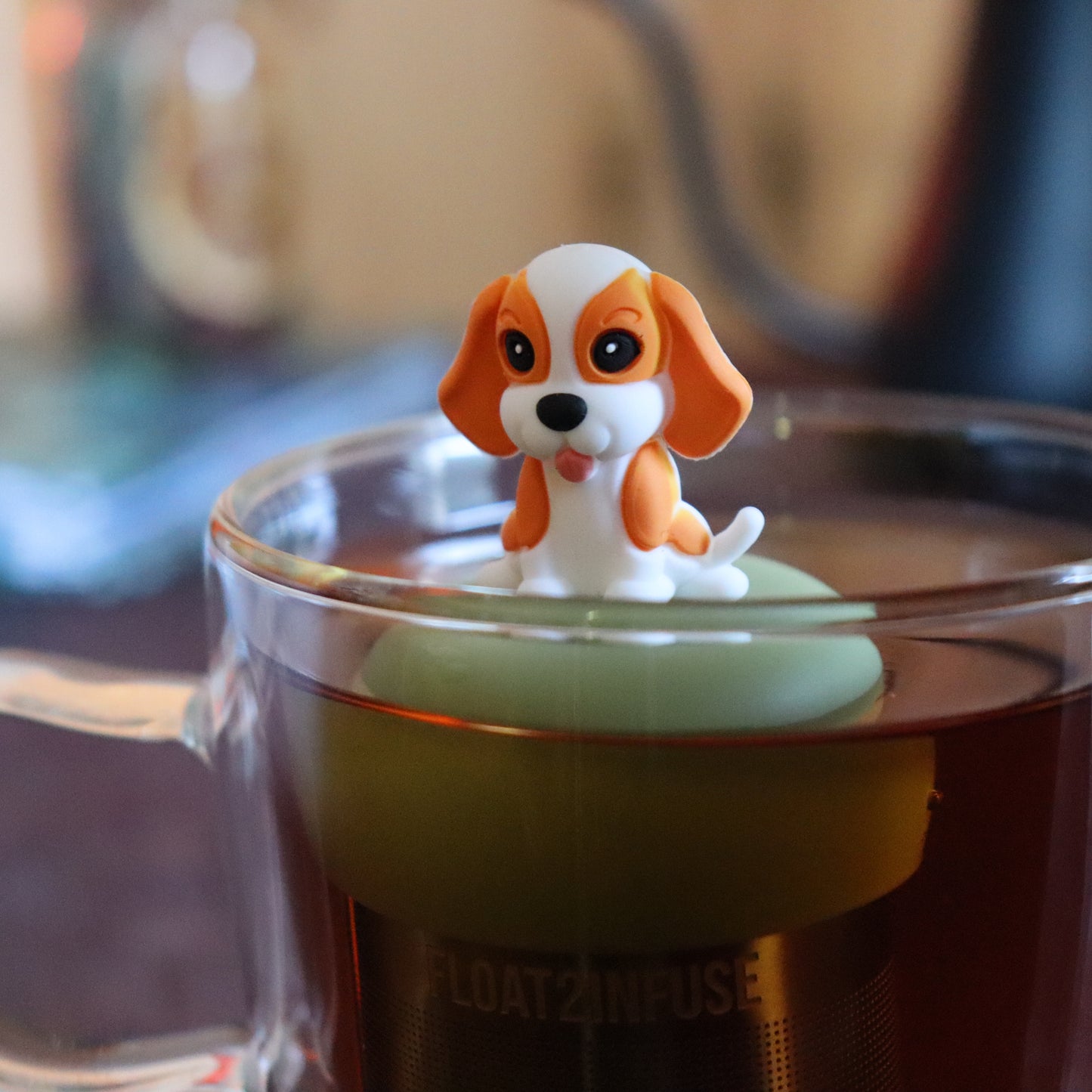 orange and white beagle animal tea infuser for loose leaf tea with a fine mesh stainless steel tea strainer that reads FLOAT2INFUSE on a green base floating in a glass tea mug with tea kettle in the background
