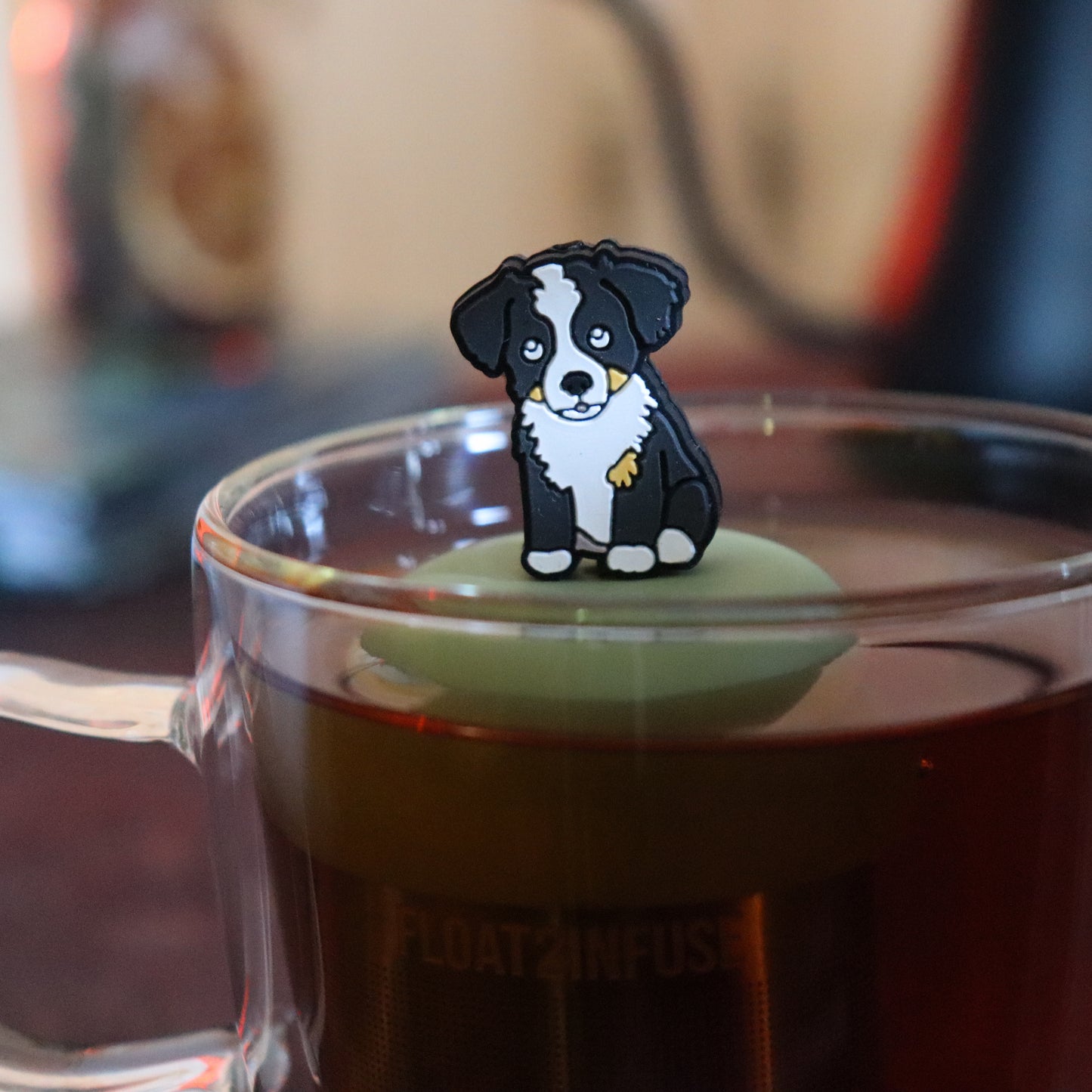 black and white border collie dog animal tea infuser for loose leaf tea with a fine mesh stainless steel tea strainer that reads FLOAT2INFUSE on a green base floating in a glass tea mug with tea kettle in the background