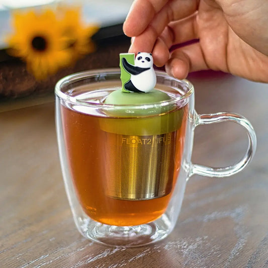 black and white panda climbing green bamboo for loose leaf tea with fine mesh stainless steel tea strainer that reads FLOAT2INFUSE on a green base floating in a glass tea mug with flowers in the backgroun