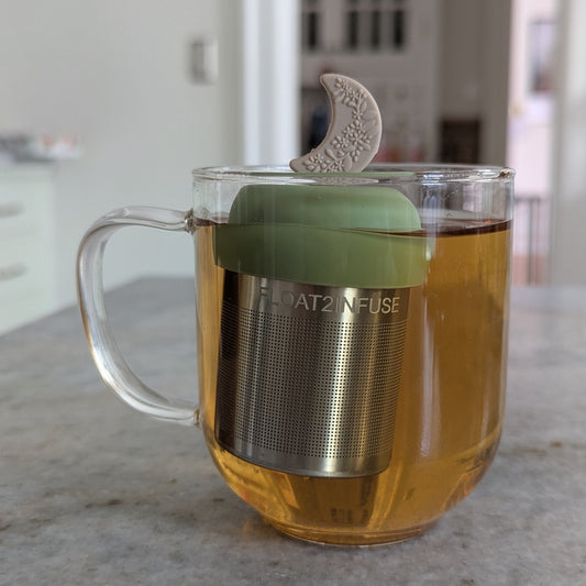 beige crescent moon with raised floral pattern tea infuser for loose leaf tea with fine mesh stainless steel tea strainer that reads FLOAT2INFUSE on a green base floating in a glass tea mug