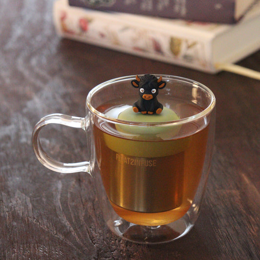 black baby highland cow animal tea infuser for loose leaf tea on a green base with a fine mesh stainless steel tea infuser that reads FLOAT2INFUSE floating in a glass tea mug with books in the background