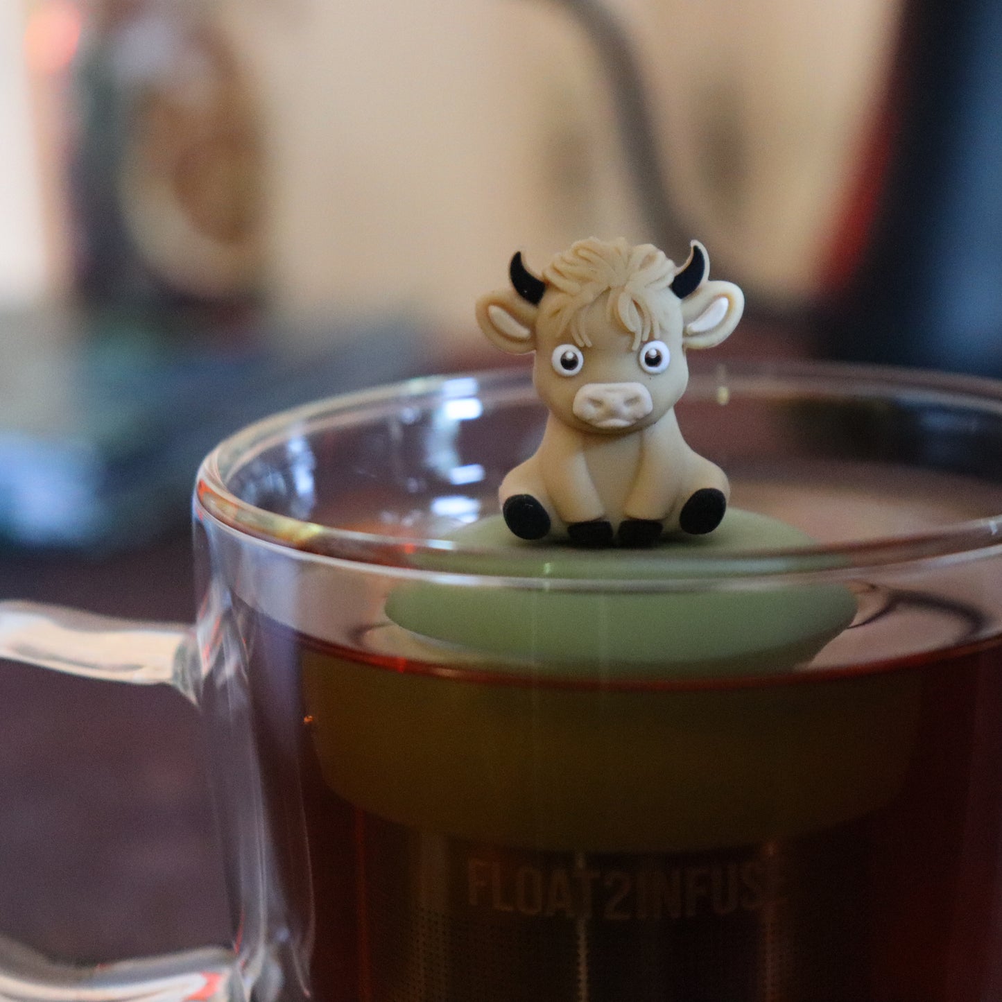 blonde baby highland cow animal tea infuser for loose leaf tea with a fine mesh stainless steel tea strainer that reads FLOAT2INFUSE on a green base floating in a glass tea mug with tea kettle in the background