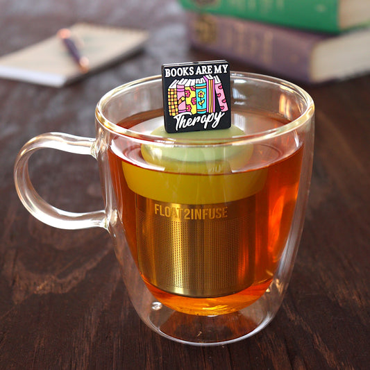 black square with text that reads "books are my therapy" on books tea infuser for loose leaf tea with a fine mesh stainless steel tea infuser that reads FLOAT2INFUSE on a green base floating in a glass tea mug with pen on notebook and books in the background