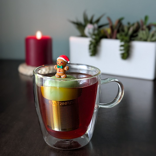 Smiling gingerbread man wearing a red santa hat and a green bowtie infuser for loose leaf tea with a fine mesh stainless steel tea strainer that reads FLOAT2INFUSE on a green base with a lit candle and plants in the background