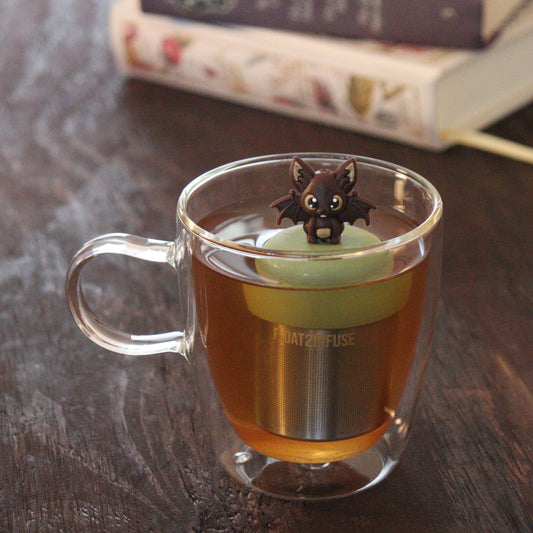 brown bat animal tea infuser for loose leaf tea with a fine mesh stainless steel tea infuser that reads FLOAT2INFUSE on a green base floating in a glass tea mug with books in the background
