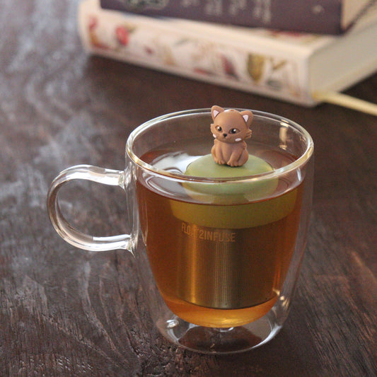brown cat animal tea infuser for loose leaf tea on a green base with a fine mesh stainless steel tea infuser that reads FLOAT2INFUSE floating in a glass tea mug with books in the background