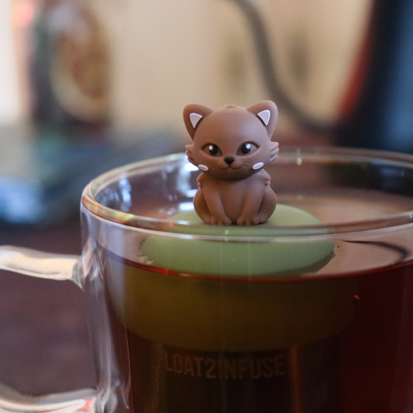 brown cat animal tea infuser for loose leaf tea with a fine mesh stainless steel tea strainer that reads FLOAT2INFUSE on a green base floating in a glass tea mug with tea kettle in the background