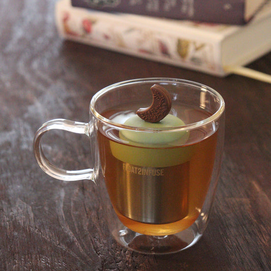 brown floral crescent moon tea infuser for loose leaf tea with a fine mesh stainless steel tea infuser that reads FLOAT2INFUSE on a green base floating in a glass tea mug with books in the background