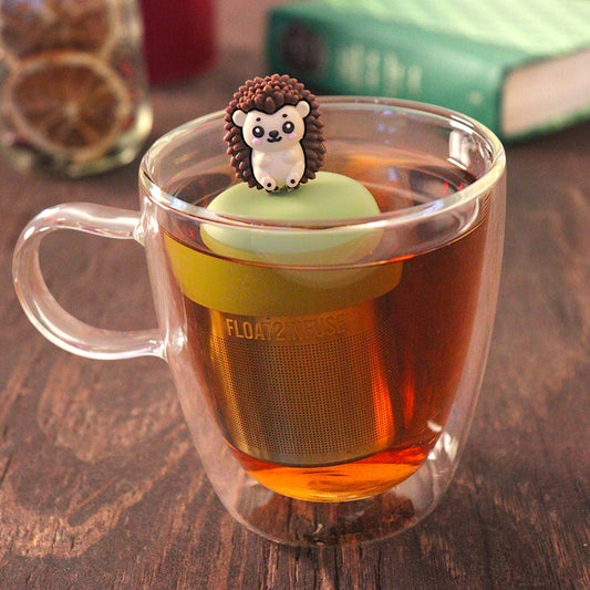 brown hedgehog tea infuser for loose leaf tea with a fine mesh stainless steel tea infuser that reads FLOAT2INFUSE on a green base floating in a glass tea mug with loose leaf tea and books in the background