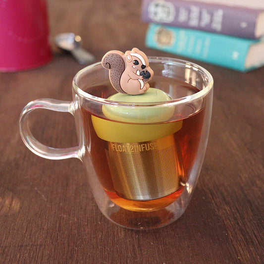 Brown squirrel holding an acorn tea infuser for loose leaf tea with a fine mesh stainless steel tea strainer
that reads FLOAT2INFUSE on a green base with a tea canister and books in the background