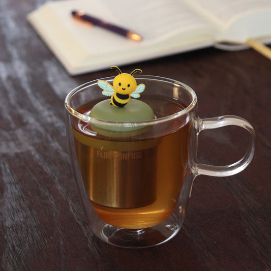yellow and black bee animal tea infuser for loose leaf tea with a fine mesh stainless steel tea infuser that reads FLOAT2INFUSE on a green base floating in a glass tea mug with notebook and pen in the background