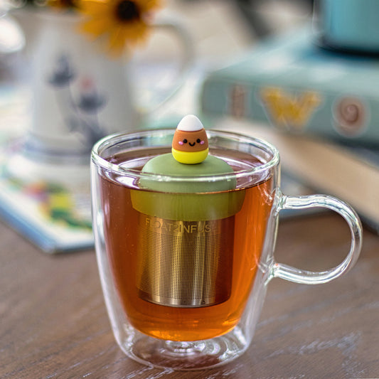 candy corn with a smiling face tea infuser for loose leaf tea with a fine mesh stainless steel tea strainer that reads FLOAT2INFUSE on a green base floating in a glass tea mug
with books in the background