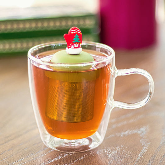red mitten with green christmas tree and white accents tea infuser for loose leaf tea with a fine mesh stainless steel tea strainer that reads FLOAT2INFUSE on a green base floating in a glass tea mug
with books in the background