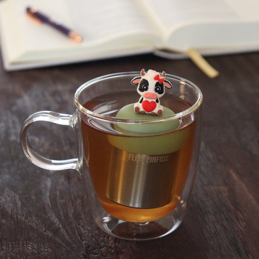 white and black cow with red bow and heart animal tea infuser for loose leaf tea with a fine mesh stainless steel tea infuser that reads FLOAT2INFUSE on a green base floating in a glass tea mug with notebook and pen in the background