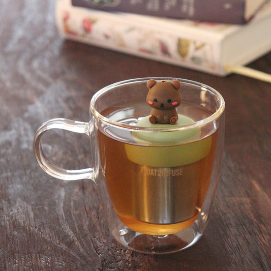 blushing brown bear animal tea infuser for loose leaf tea with a fine mesh stainless steel tea infuser that reads FLOAT2INFUSE on a green base floating in a glass tea mug with books in the background
