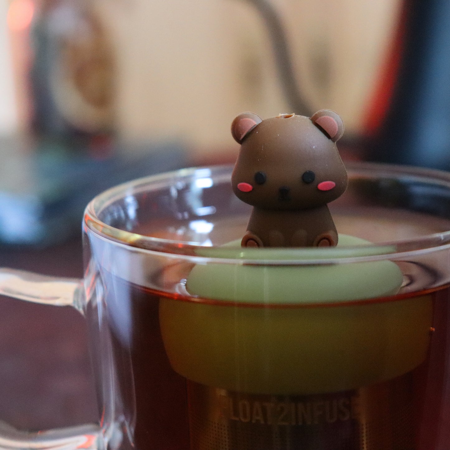 cute brown blushing bear animal tea infuser for loose leaf tea with a fine mesh stainless steel tea strainer that reads FLOAT2INFUSE on a green base floating in a glass tea mug with tea kettle in the background